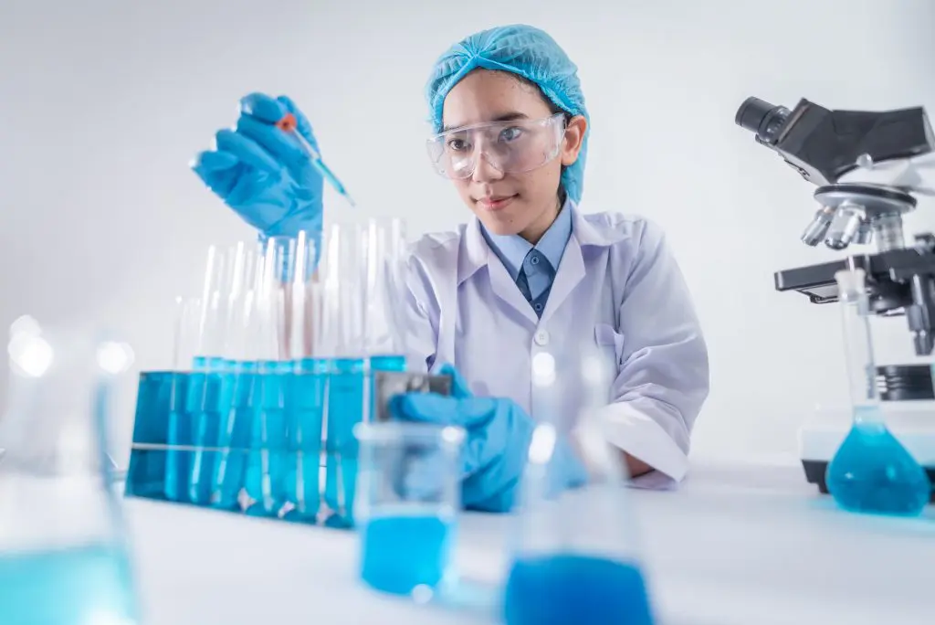 A woman in lab coat and goggles working with blue liquid.